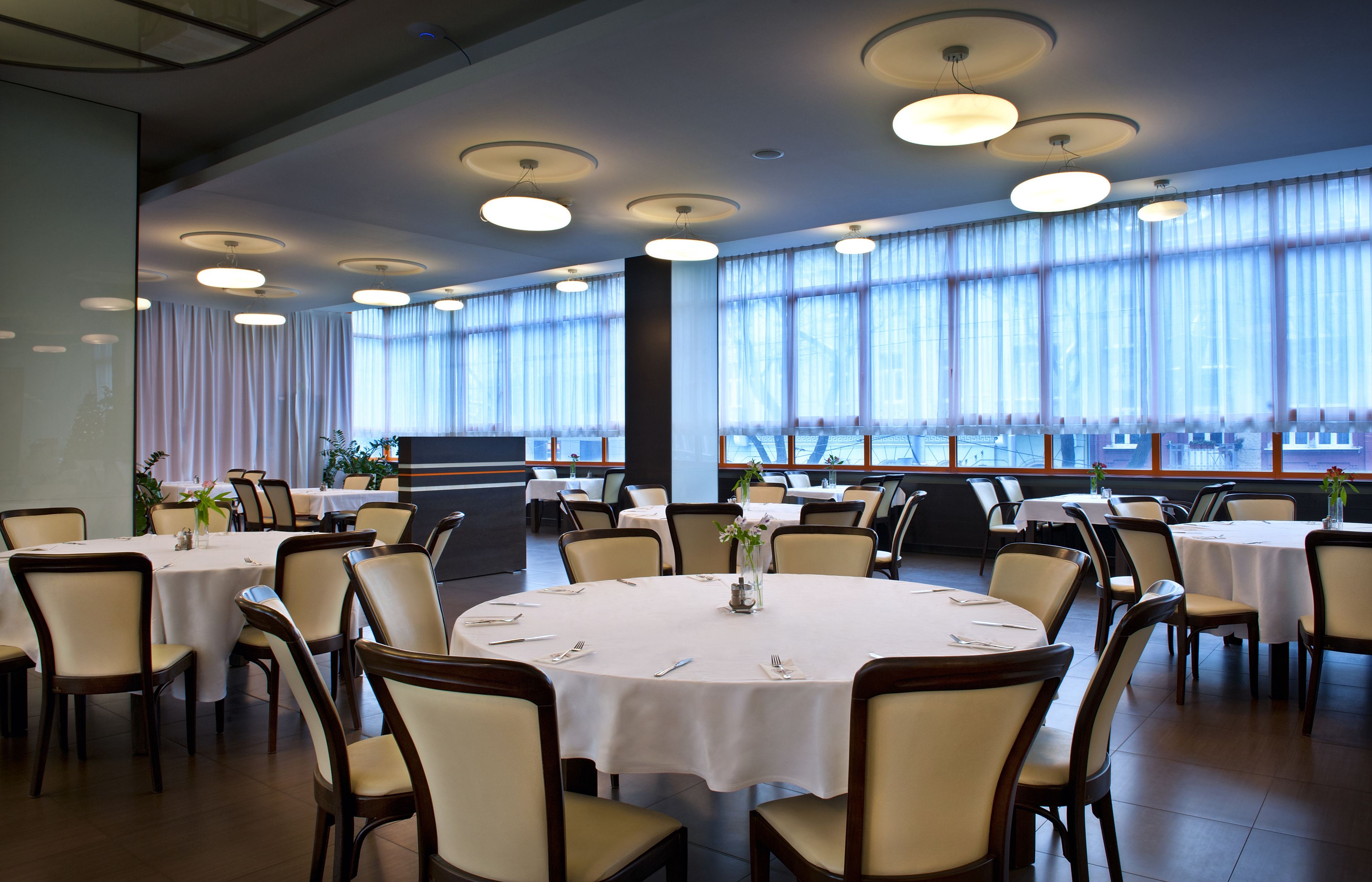 A spacious dining area with round tables and chairs, large windows with sheer curtains, and ceiling lights.