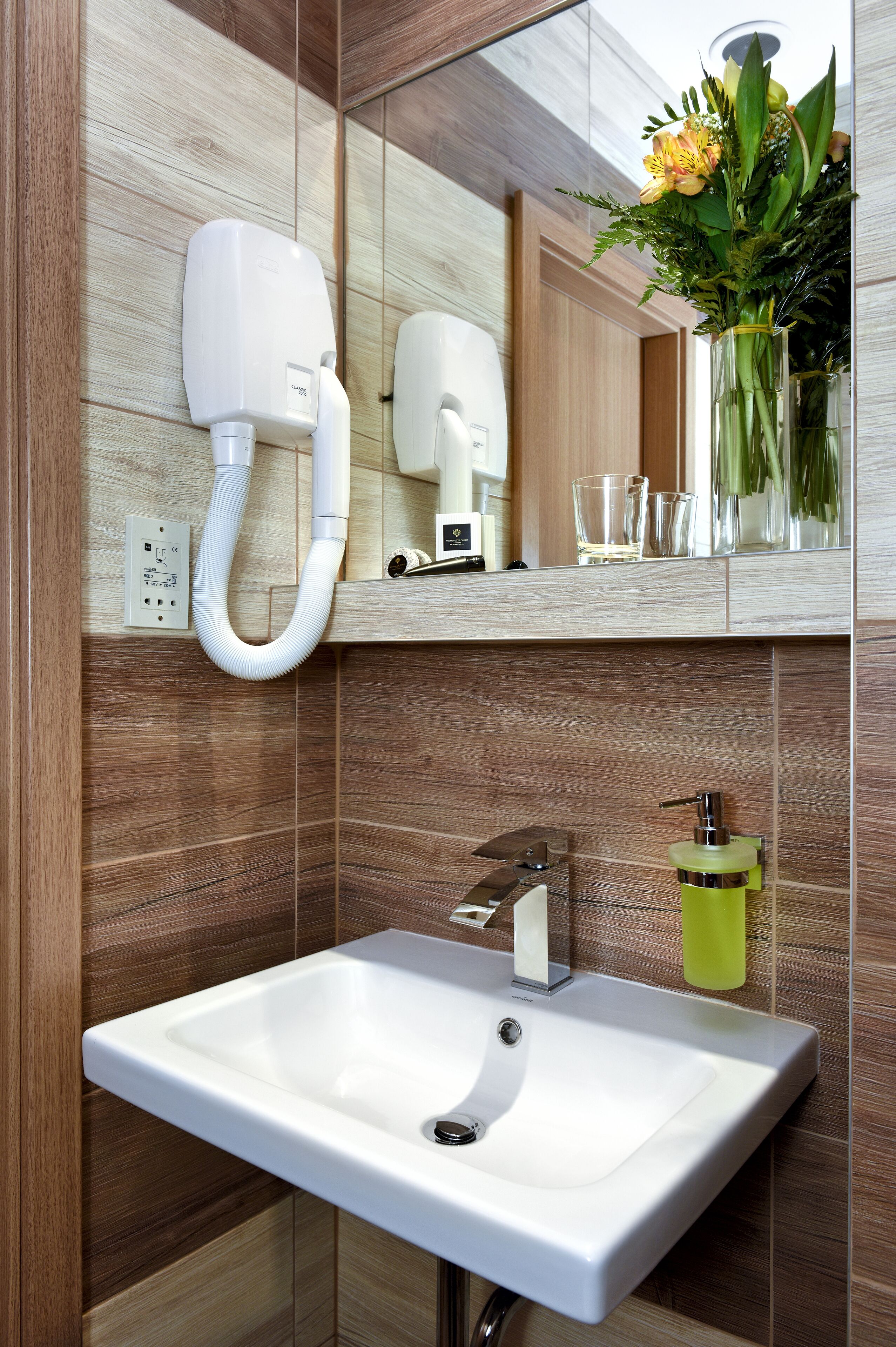 A bathroom with a white sink, a mirror, a hairdryer, and a vase with flowers.