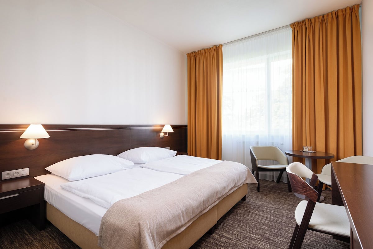 A hotel room with a double bed, two bedside lamps, a small table with chairs, and a window with curtains.
