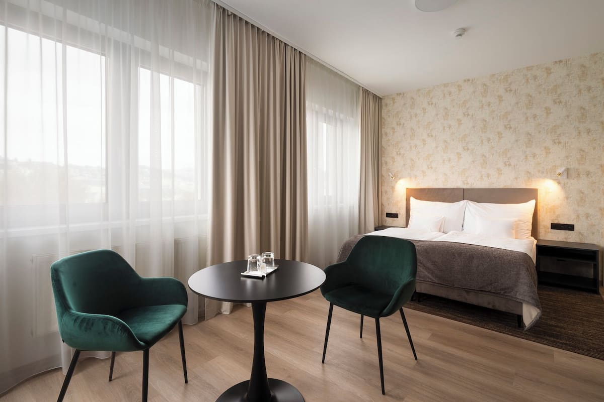A hotel room with a bed, two chairs, a table, and a view through sheer curtains.