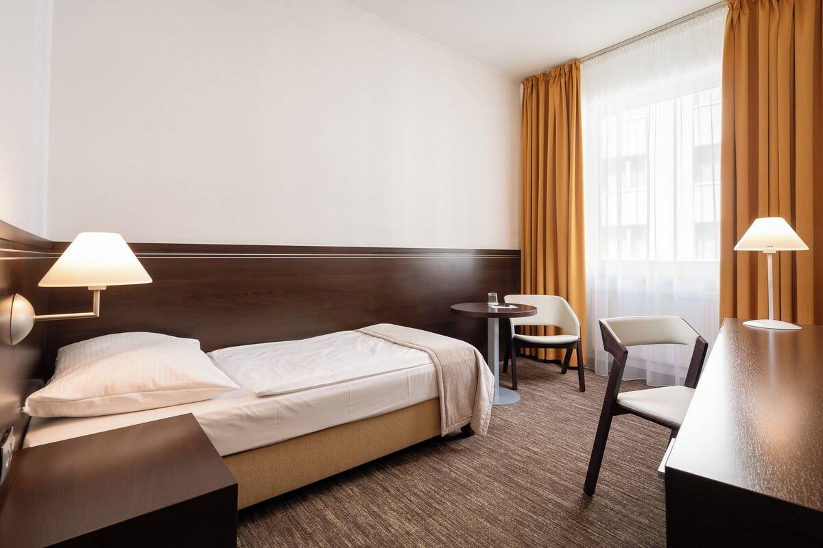 A hotel room with a bed, two bedside lamps, a small round table with a chair, and a desk with a chair.