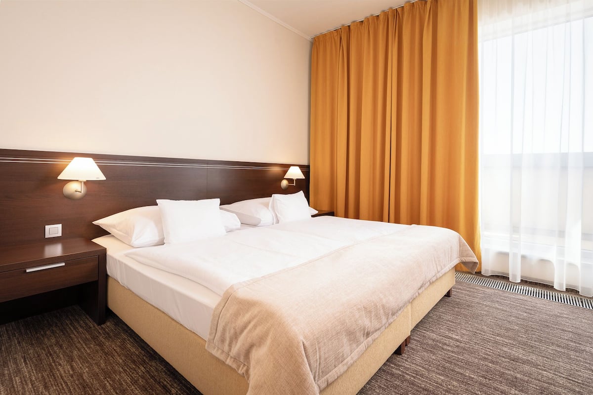 A hotel room with a bed, two bedside lamps, a nightstand, a window with curtains, and a carpeted floor.