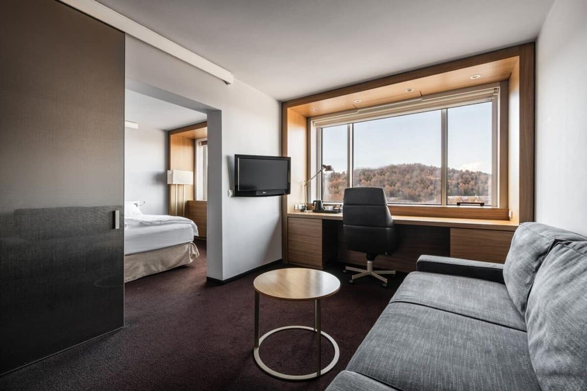 Una habitación de hotel moderna con cama, escritorio con silla, televisor y un ventanal que ofrece vistas al exterior.