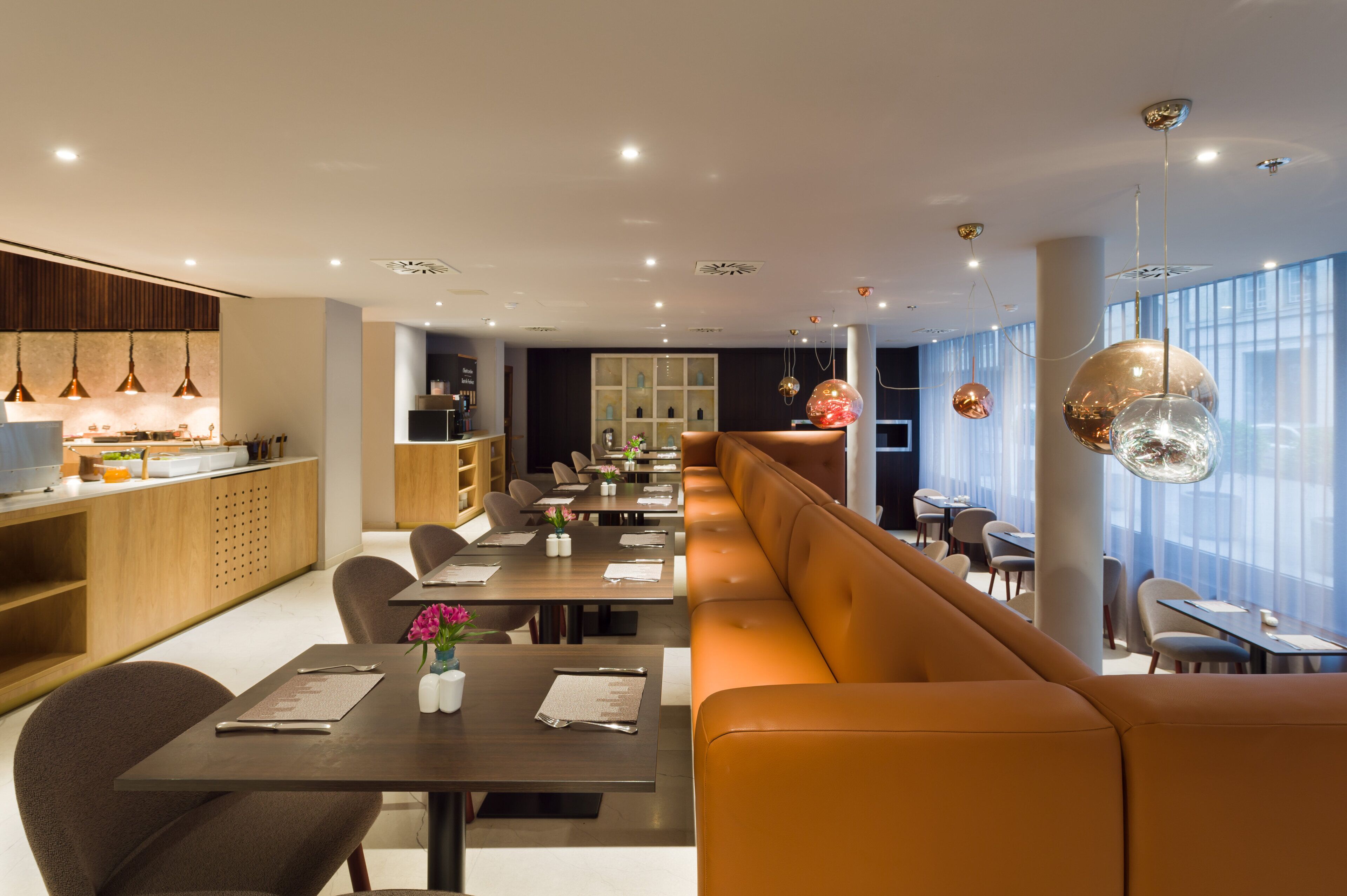 A modern restaurant with long communal tables, cushioned seating, and pendant lighting.