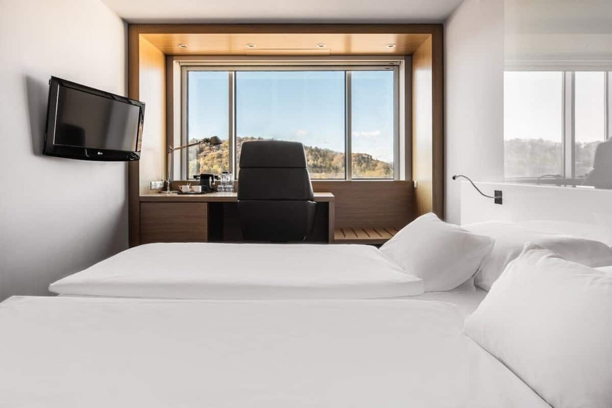 Habitación de hotel moderna con una cama grande, un escritorio con silla, televisor montado en la pared y un ventanal con vistas al exterior.