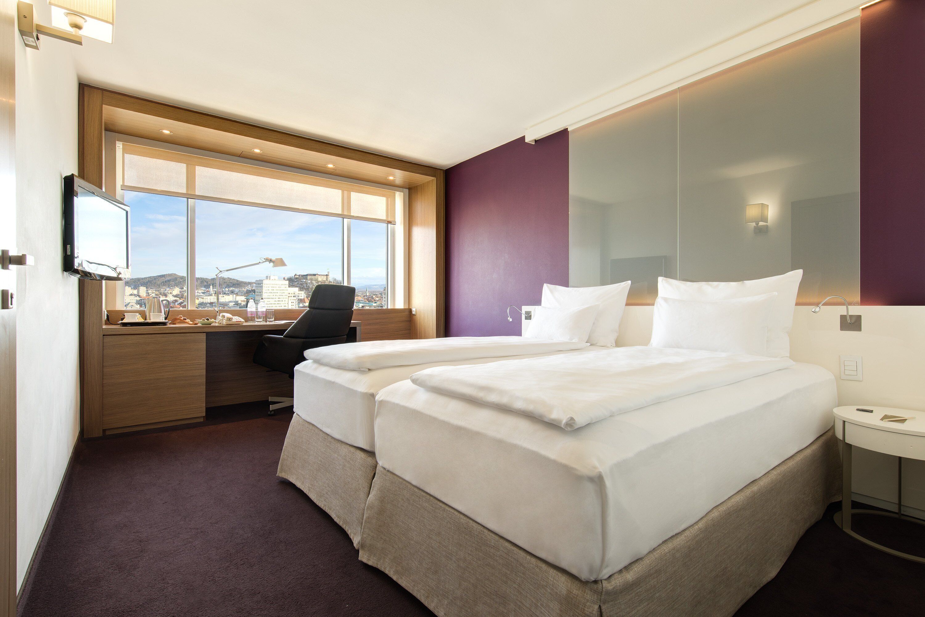 Habitación de hotel con dos camas, un escritorio y un ventanal grande con vista a la ciudad.