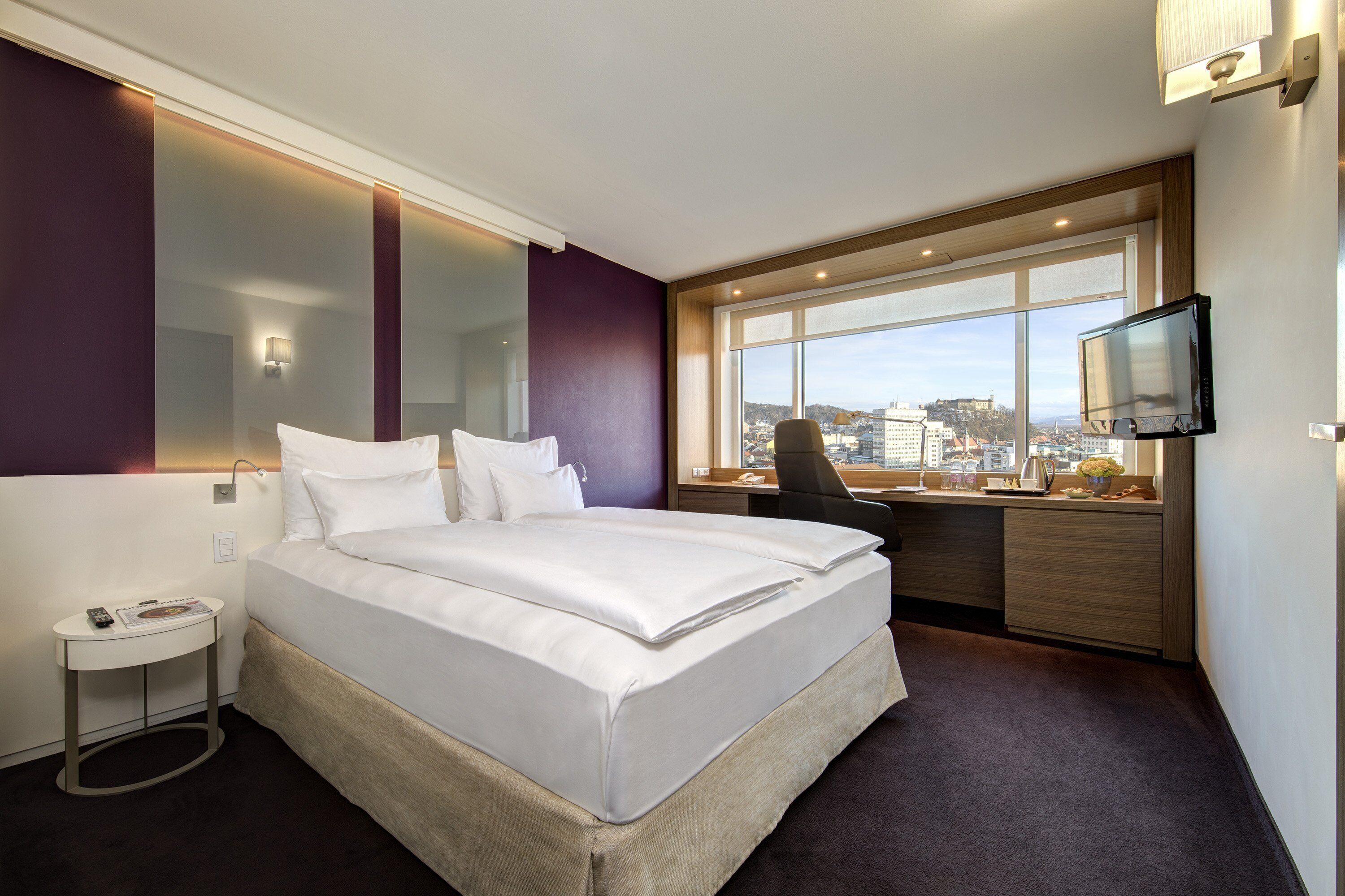 Ein modernes Hotelzimmer mit einem großen Bett, einem Schreibtisch, einem Sessel, einem Fernseher und Blick auf die Stadt.