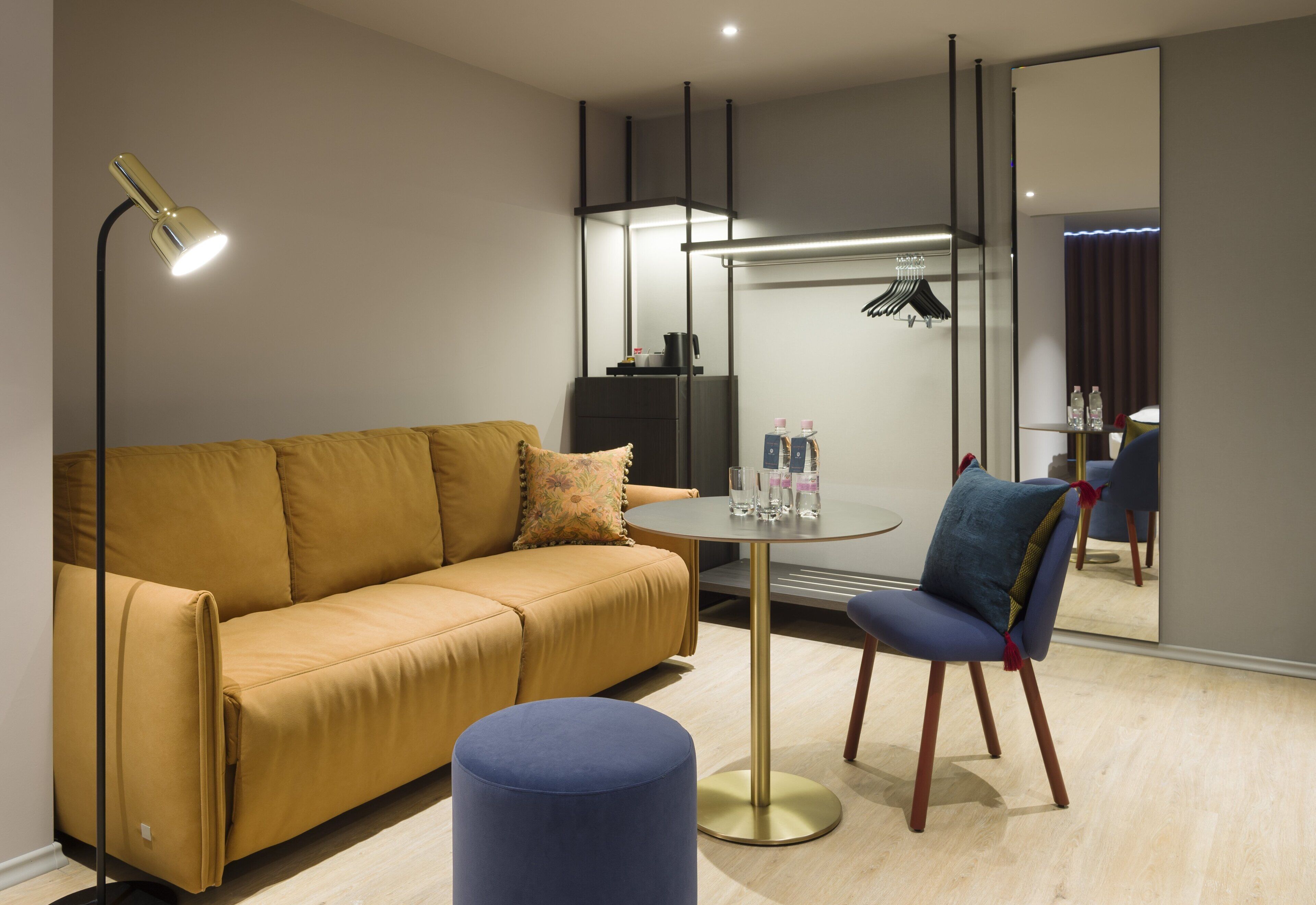 Una habitación de hotel moderna con sofá, una mesa redonda y una silla. Hay un espejo en la pared y una lámpara en el suelo.