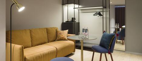 Una habitación de hotel moderna con sofá, una mesa redonda y una silla. Hay un espejo en la pared y una lámpara en el suelo.
