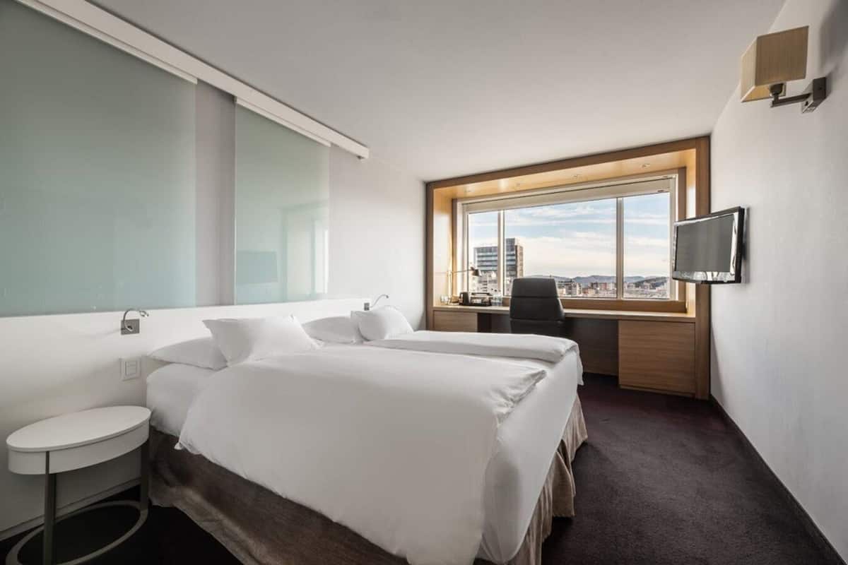 Habitación de hotel con dos camas, un escritorio, un televisor y una ventana con vista a la ciudad.