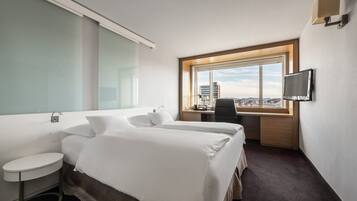 A hotel room with two beds, a desk, a TV, and a window with a city view.