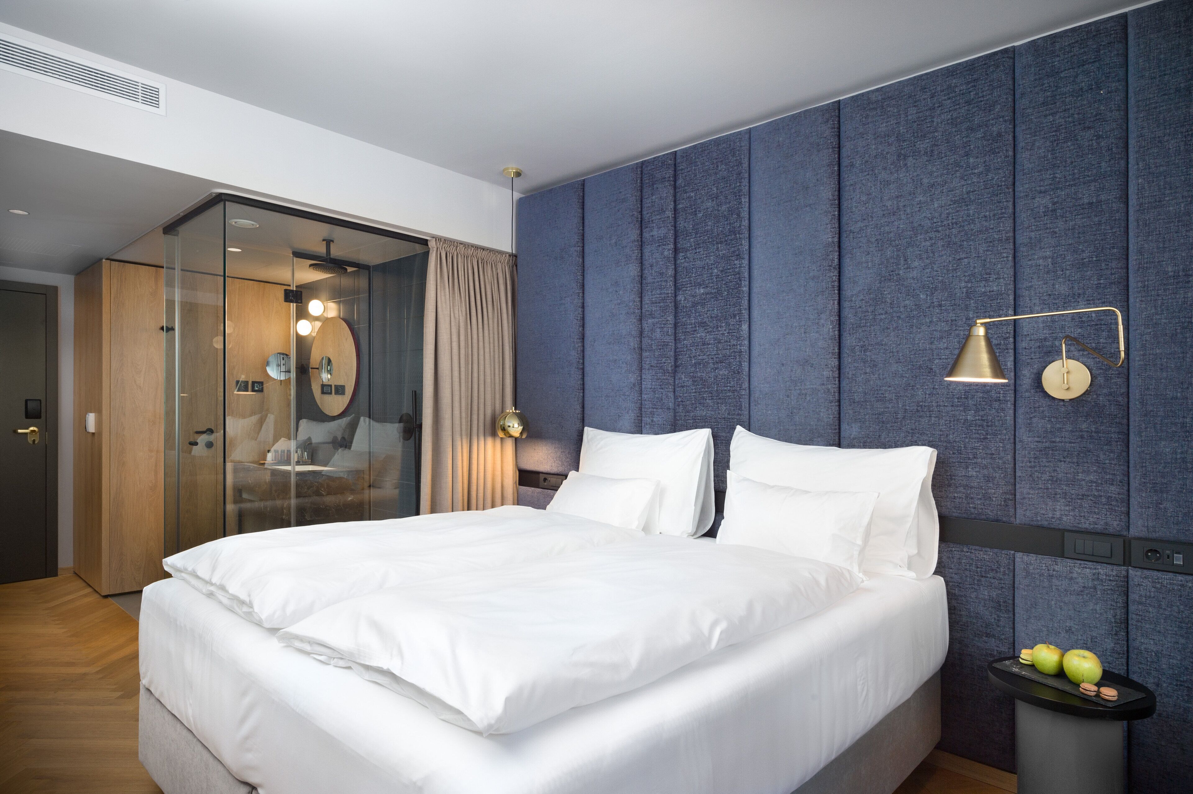 A modern hotel room with a large bed, a glass-enclosed walk-in closet, a blue textured wall, and a small table with apples and cookies.