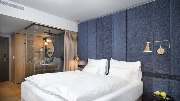 A modern hotel room with a large bed, a glass-enclosed walk-in closet, a blue textured wall, and a small table with apples and cookies. - Exe Lev (Ljubljana)
