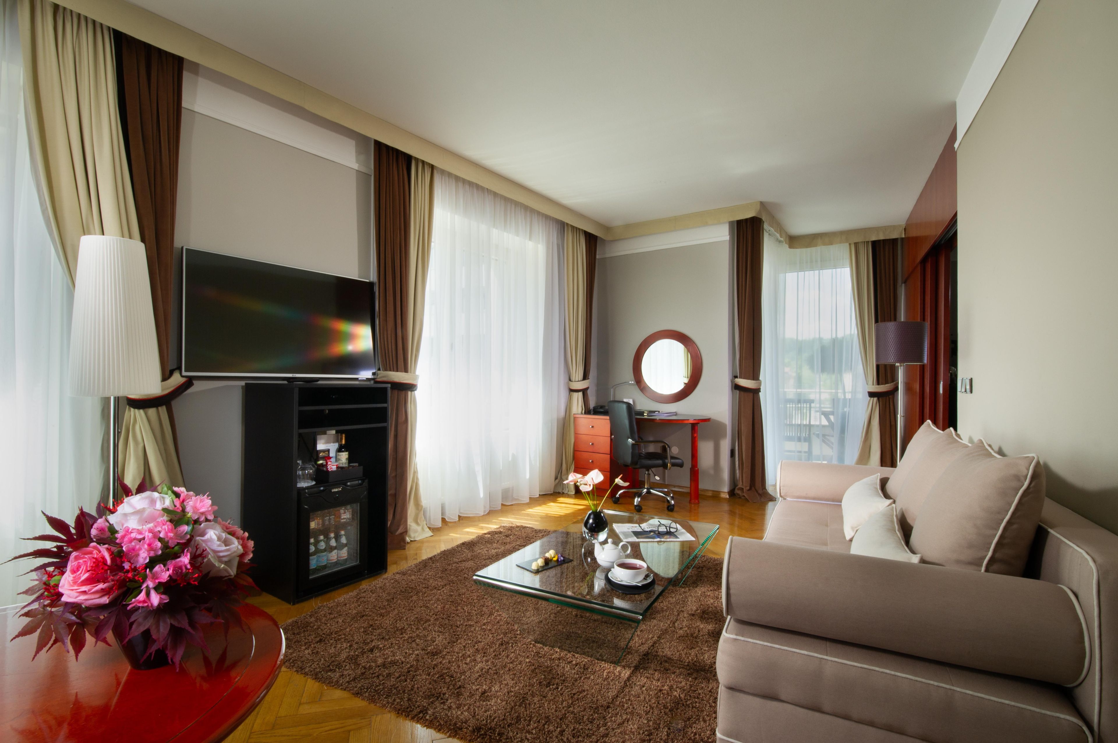 Een hotelkamer met een bank, een televisie, een klein tafeltje met een bloevas, een koelkast en een bureau met een stoel.