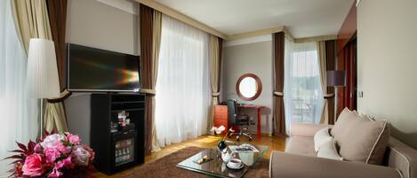 A hotel room with a sofa, a TV, a small table with a vase of flowers, a refrigerator, and a desk with a chair.