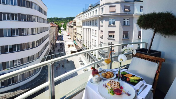 Un balcon avec une table préparée pour le petit-déjeuner, donnant sur une rue de la ville, avec des immeubles et un ciel dégagé.