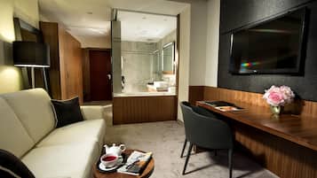Una habitación de hotel moderna con sofá, una mesa de comedor con un jarrón de flores, un televisor montado en la pared y un baño visible a través de una puerta abierta.
