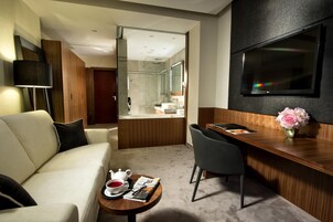Una habitación de hotel moderna con sofá, una mesa de comedor con un jarrón de flores, un televisor montado en la pared y un baño visible a través de una puerta abierta.