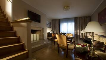 A hotel room with a staircase, a flat-screen TV mounted on the wall, a dining area with a table and chairs, a sofa, and a lamp.