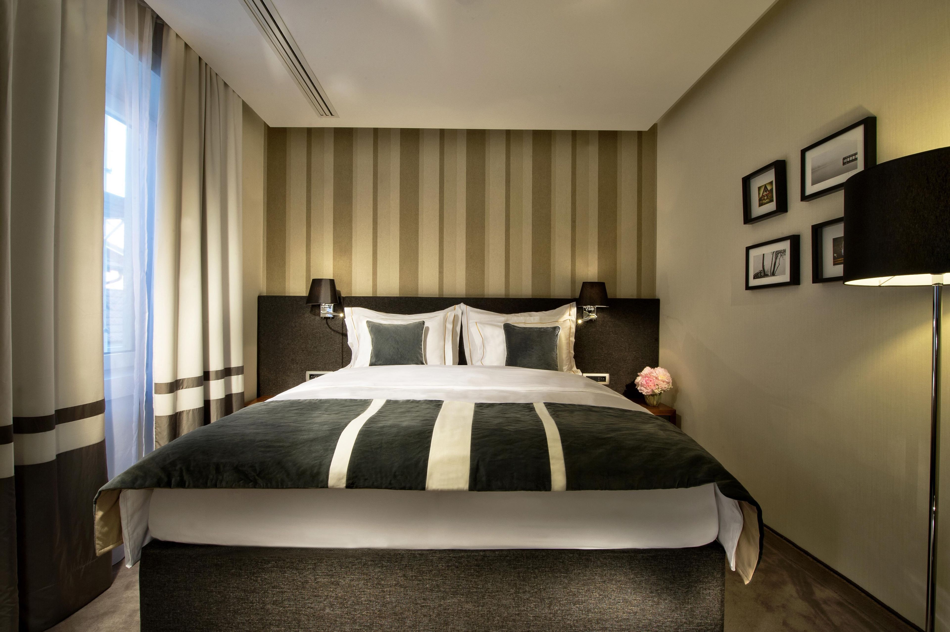 Una camera d'albergo con un letto grande, una testiera, un comodino con una lampada, una TV a muro e una finestra con tende.