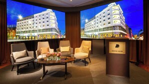 Un hall d’hôtel doté d’un grand écran affichant une image d’un bâtiment, de sièges confortables et d’un comptoir d’accueil.