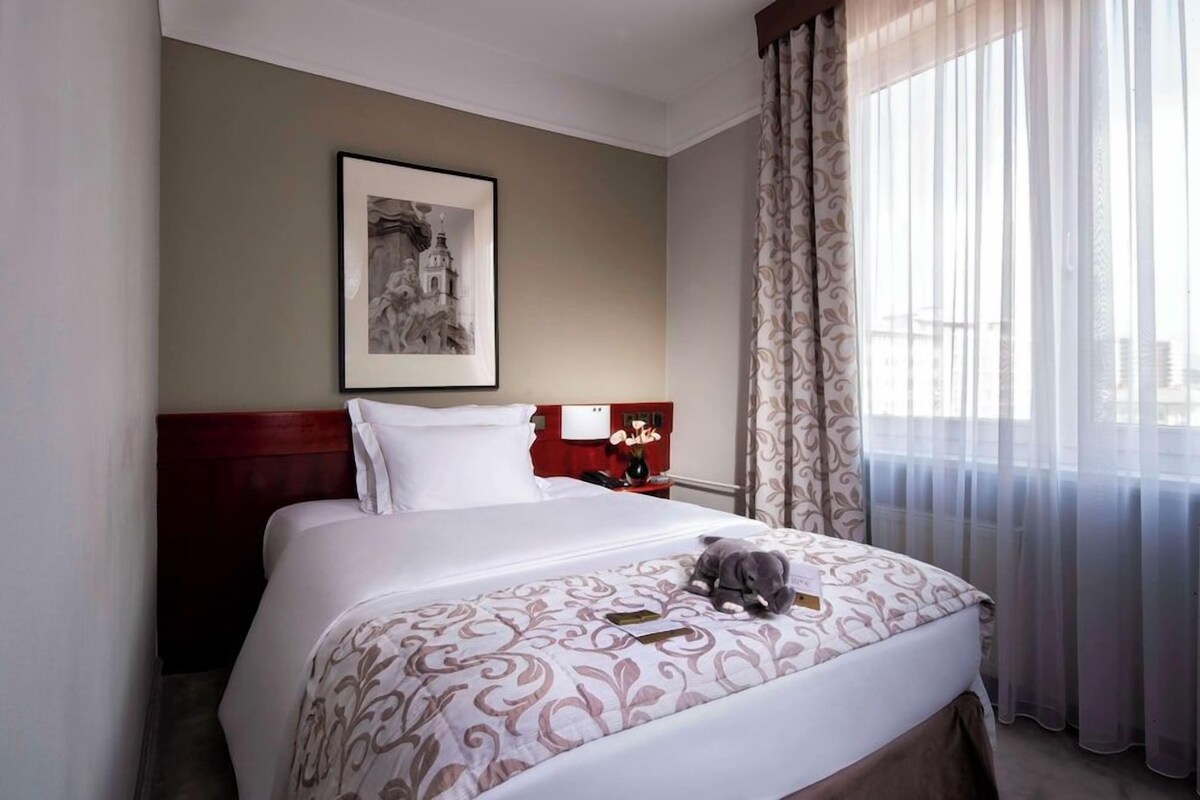 Een hotelkamer met een bed, een ingelijst beeld aan de muur, een nachtkastje met een vaas, en dunne gordijnen voor het raam.