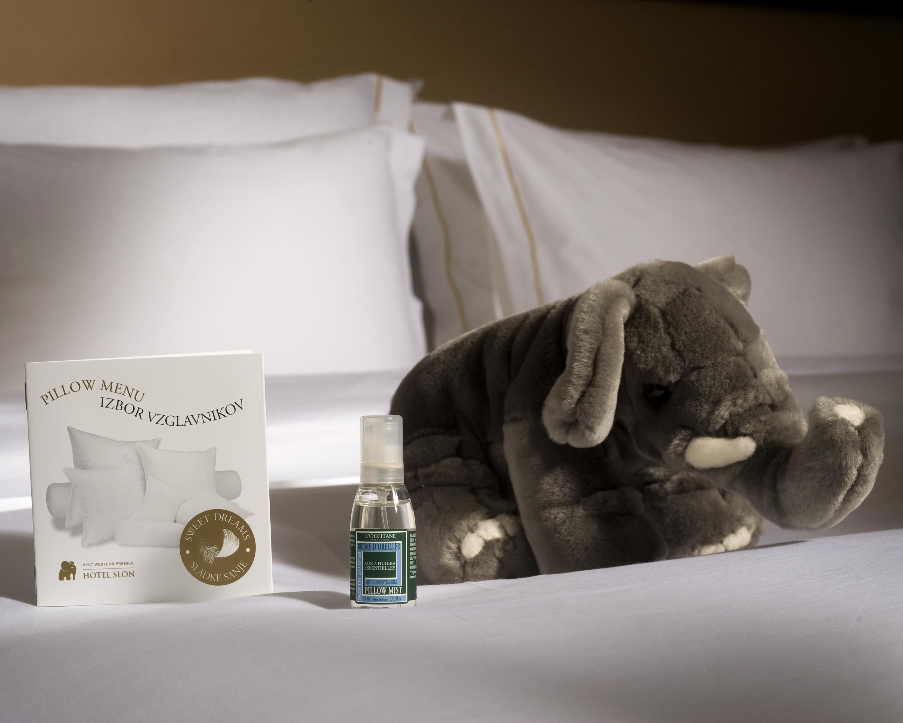 A bed with a pillow menu, a bottle of pillow mist, and a stuffed elephant toy.