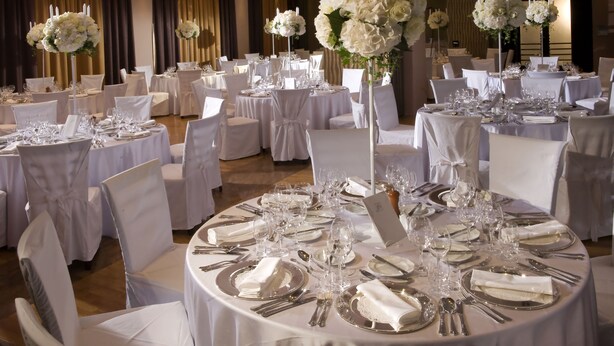 Een balzaal met ronde tafels, gedekt voor een formeel evenement, elk met witte tafelkleden, bestek en glazen.