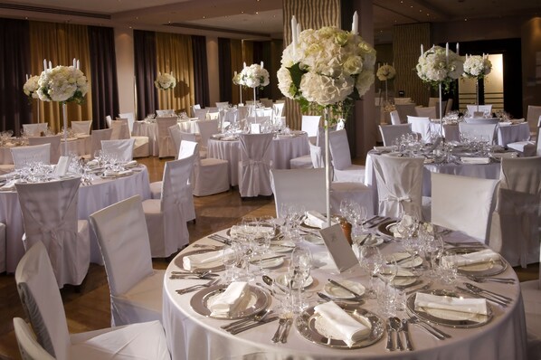 Une salle de banquet avec des tables rondes dressées pour un événement formel, chacune étant recouverte d’une nappe blanche et agrémentée de couverts en argent et de verres.