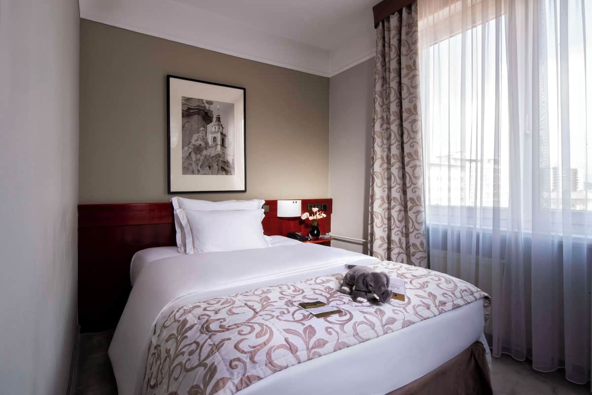 Une chambre d’hôtel avec un lit soigneusement fait, un tableau encadré au mur, une table de chevet avec une lampe et une fenêtre avec des rideaux légers.