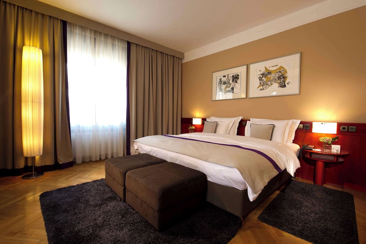Une chambre d’hôtel avec un grand lit, une table de chevet avec une lampe, une petite table avec un vase et une grande fenêtre avec des rideaux.