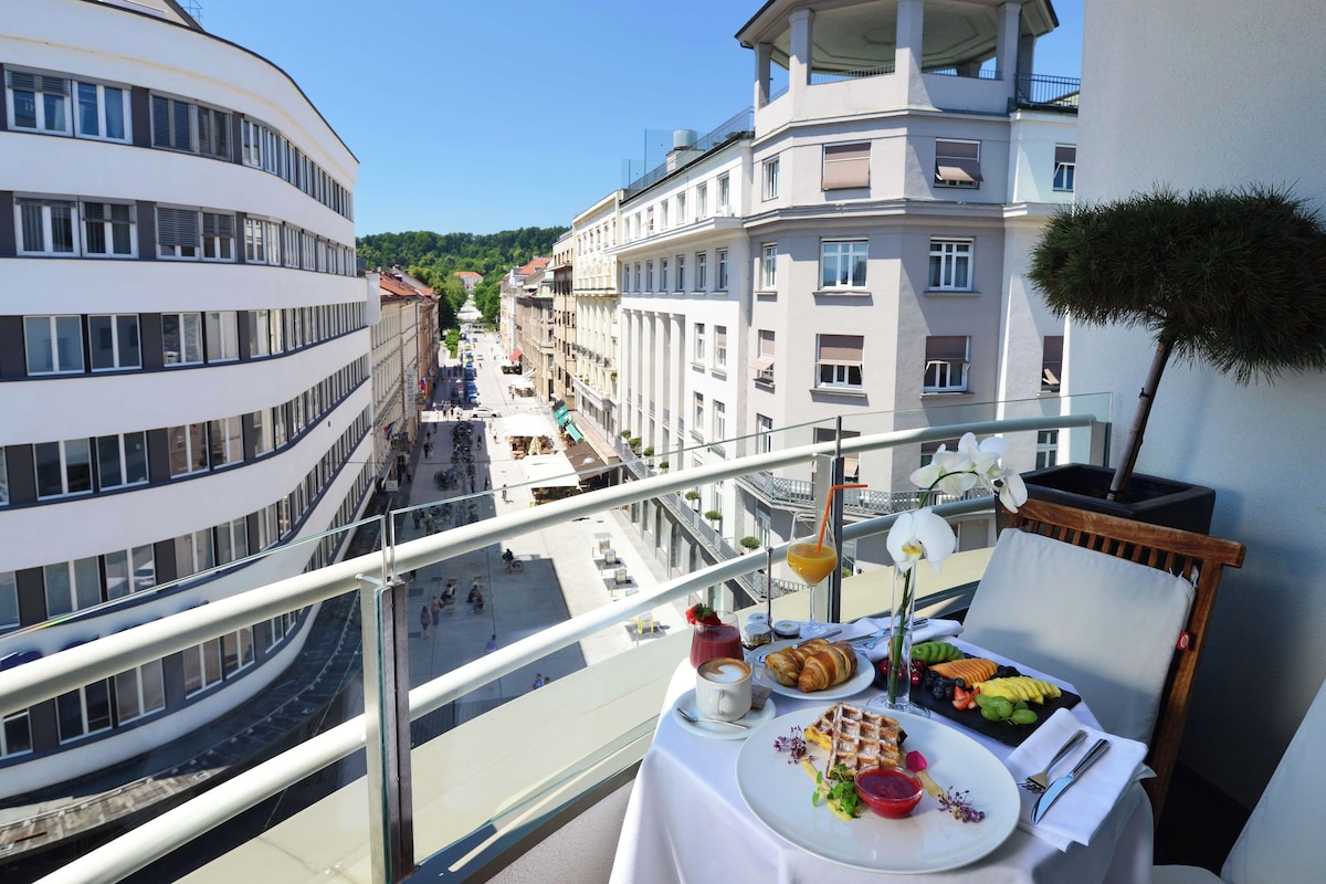 A balcony with a table set for breakfast, overlooking a city street with buildings and a clear sky.