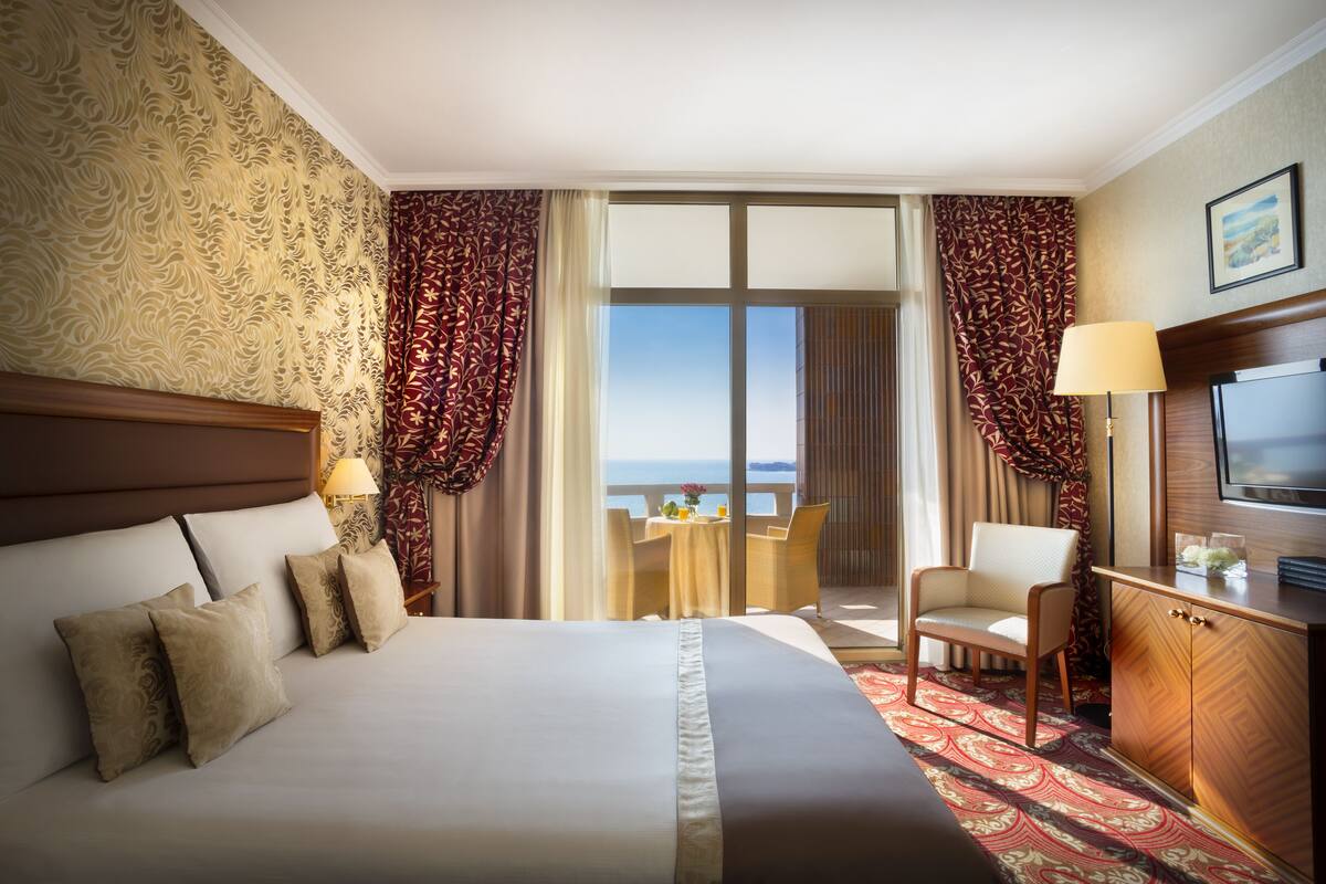 A hotel room with a large bed, a desk, a chair, a TV, and a view of the sea.