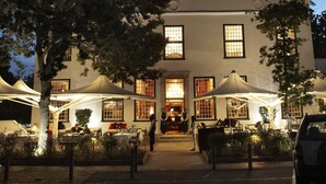 A building with a large tree in front, a parking area, and outdoor seating with umbrellas. - The Alphen Boutique Hotel & Spa (Cape Town)