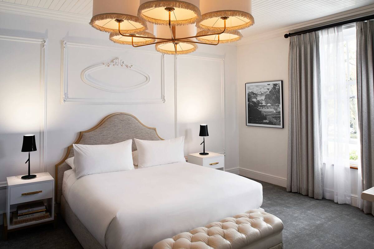 A neatly arranged bedroom with a large bed, two bedside tables, a tufted bench, a chandelier, and a framed picture on the wall.