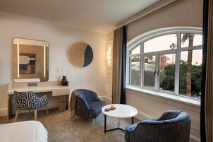 A hotel room with a large window, a desk, two armchairs, and a round table.