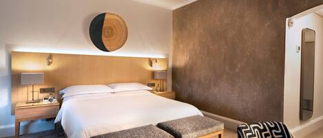 A hotel room with a large bed, bedside tables with lamps, a bench, and a patterned chair.