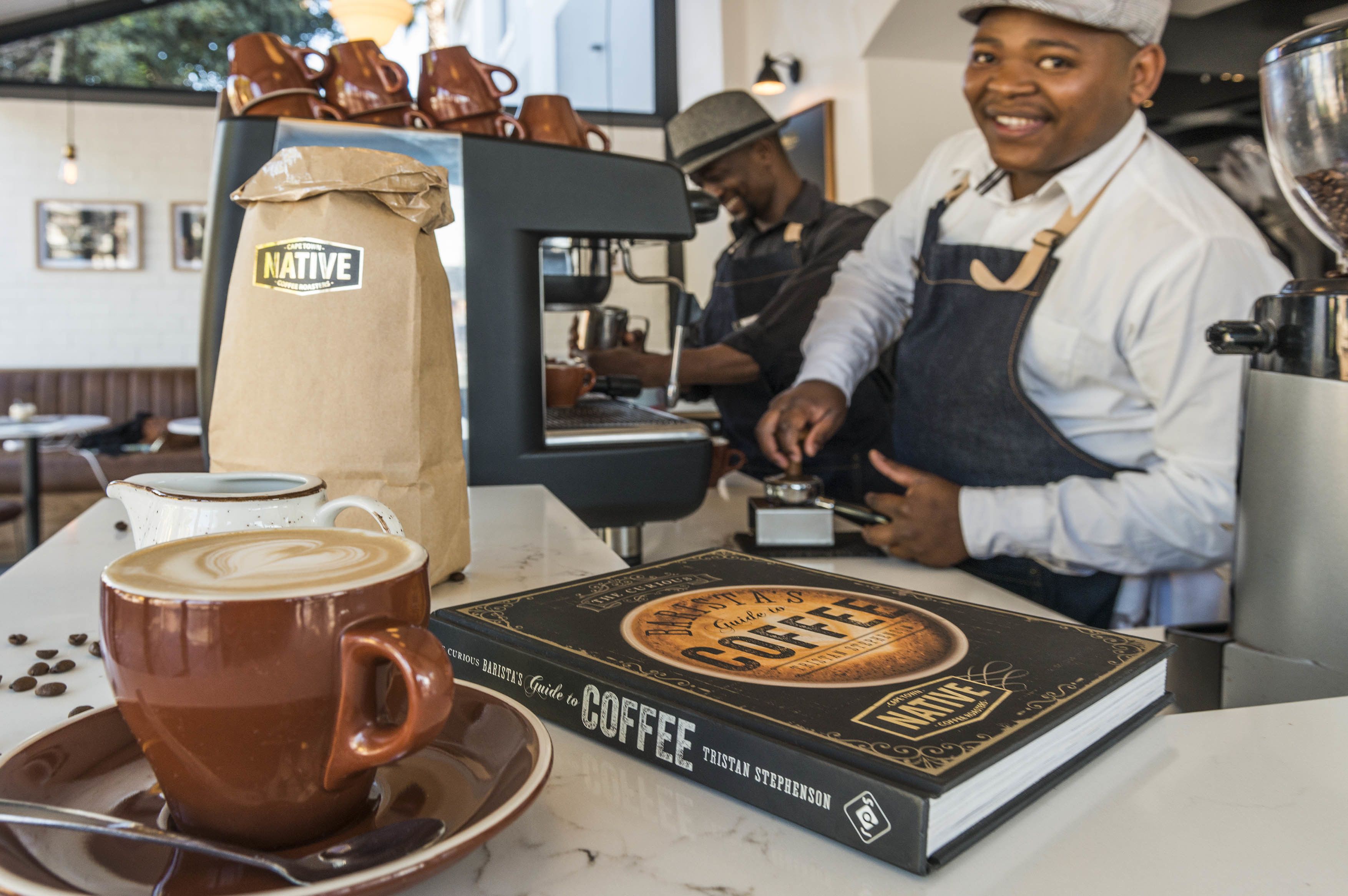 Interior de una cafetería con una máquina de café, un libro titulado 'Guide to Coffee' y una taza de café sobre el mostrador.