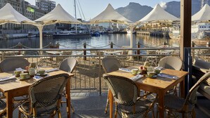 En restaurang vid vattnet med bord och stolar under parasoller, med utsikt över en småbåtshamn med båtar och en stadsbild i bakgrunden.