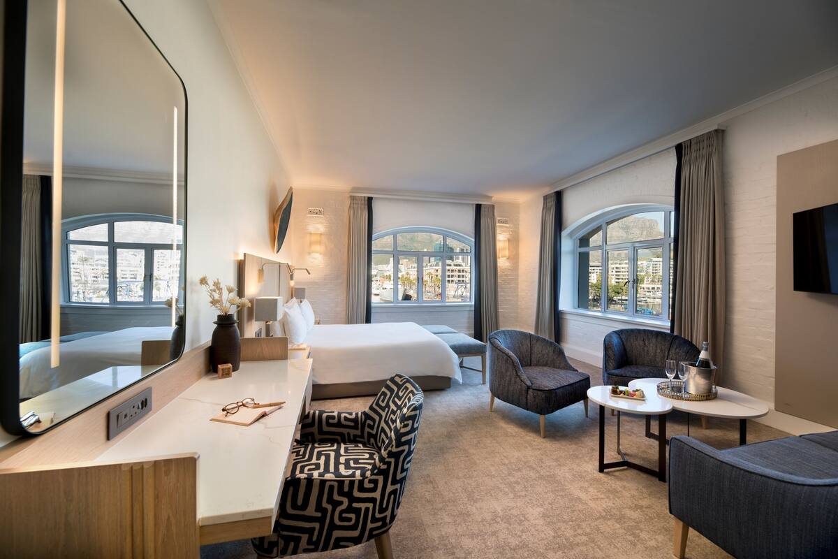 A hotel room with a large bed, a desk with a mirror, two armchairs, a small table with a tray, and large windows with curtains.