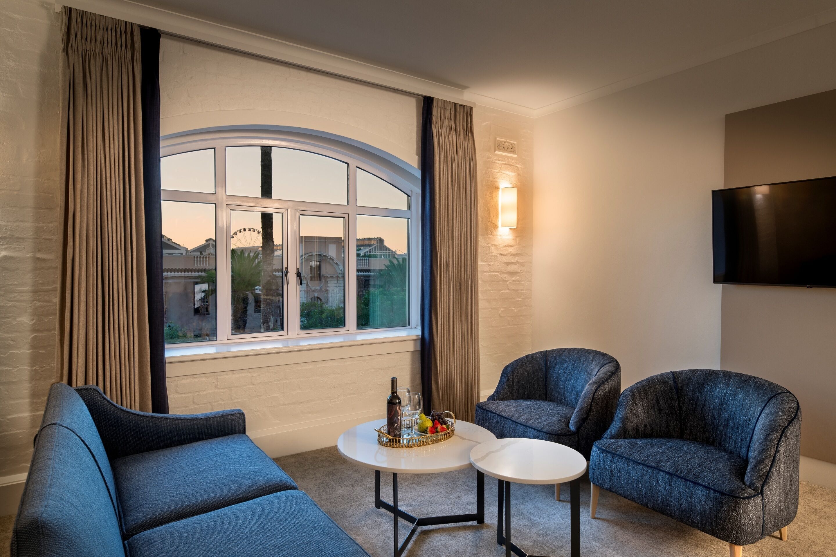 A modern living room with a curved window, two blue armchairs, a small round table with a fruit basket, and a wall-mounted TV.
