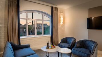 A modern living room with a curved window, two blue armchairs, a small round table with a fruit basket, and a wall-mounted TV.