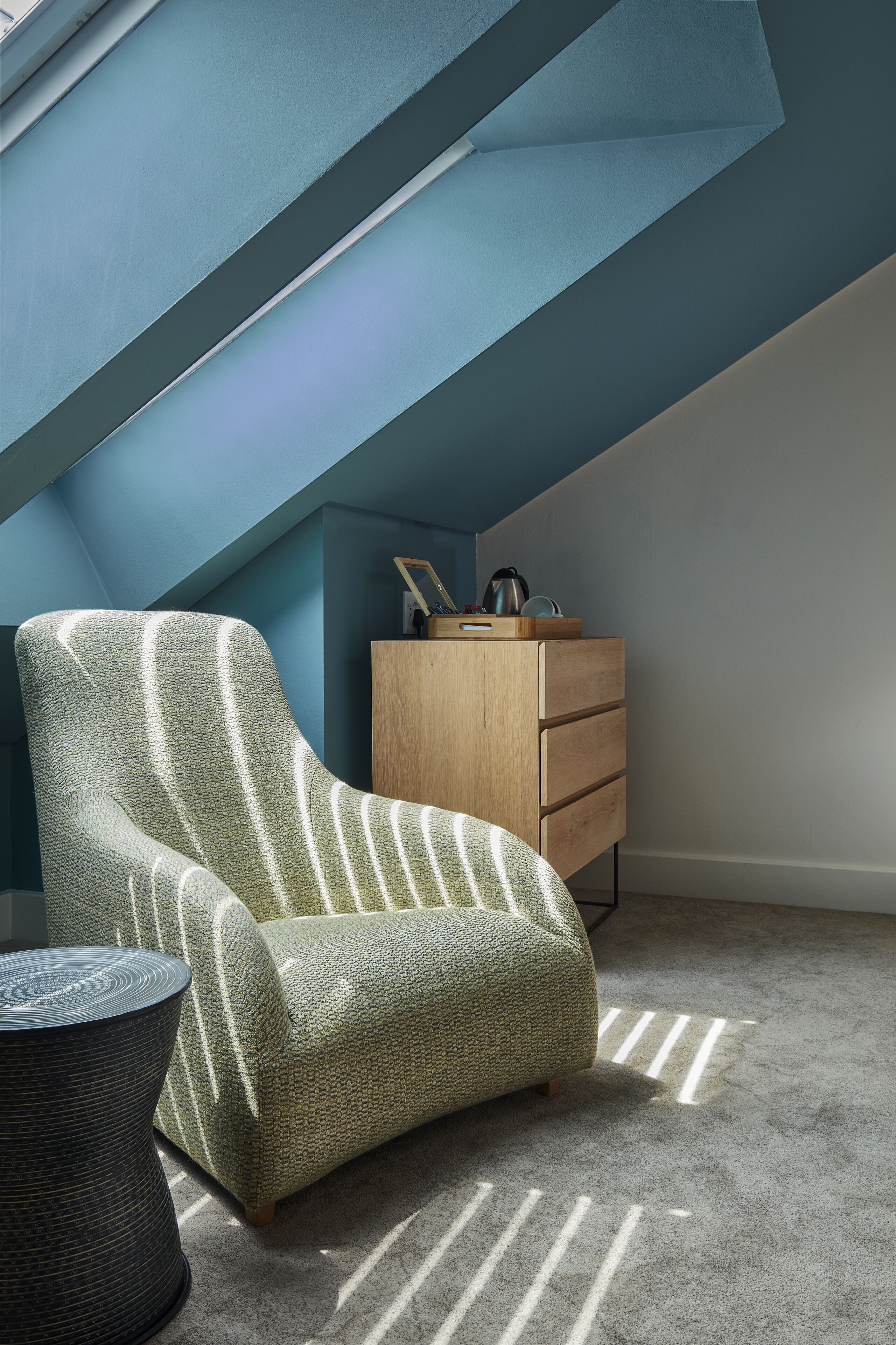 Una habitación con techo inclinado, un sillón verde a rayas, un aparador de madera y una mesa redonda.