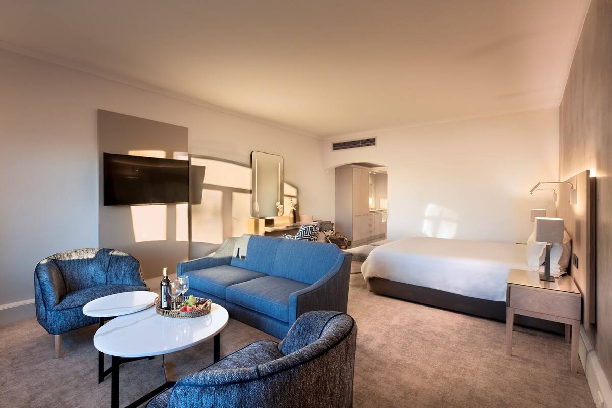 A modern hotel room with a large bed, a blue sofa, a small table with a tray of fruit, and a TV.
