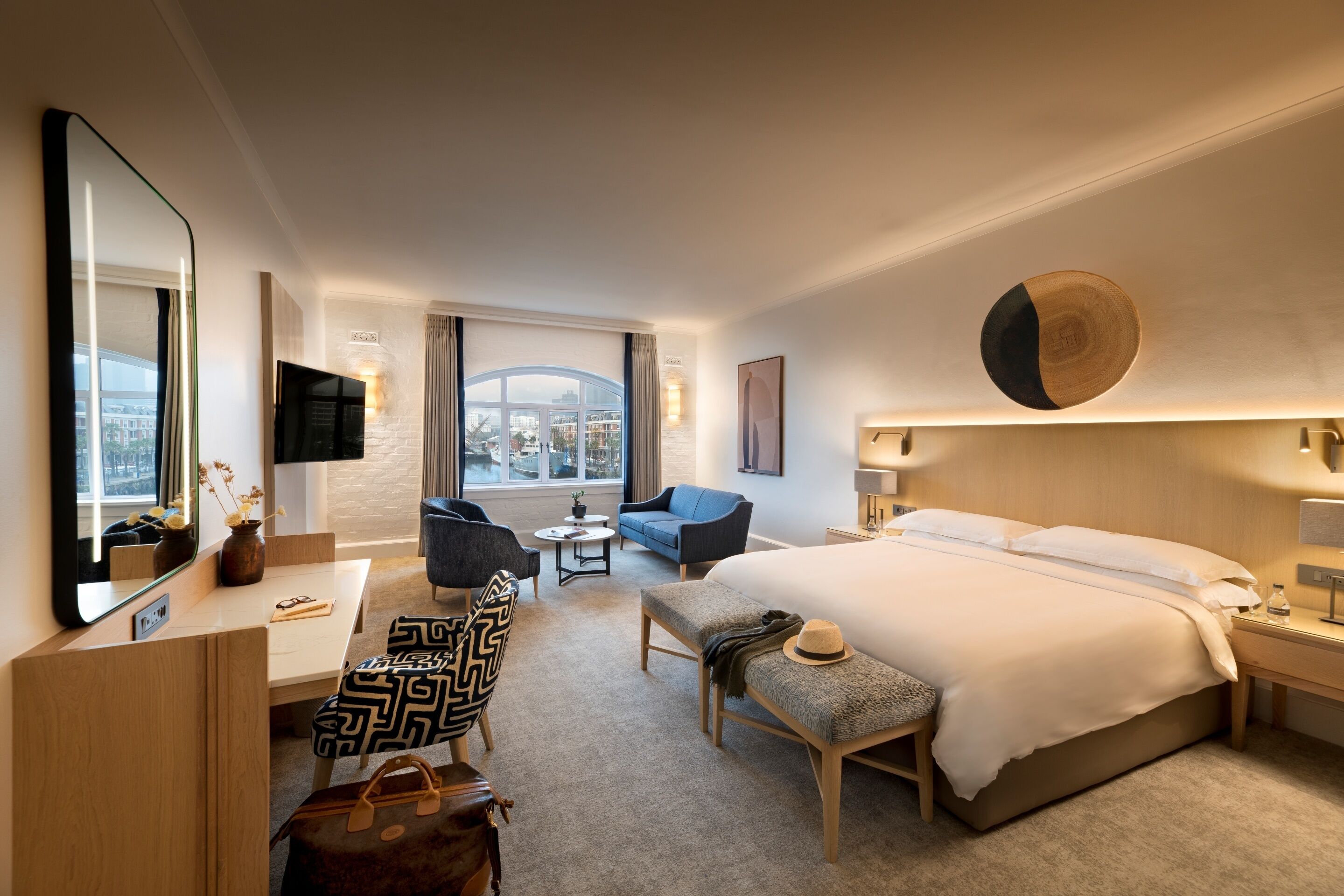 A hotel room with a large bed, a desk with a chair, a sofa, a small table, and a view of the city through large windows.