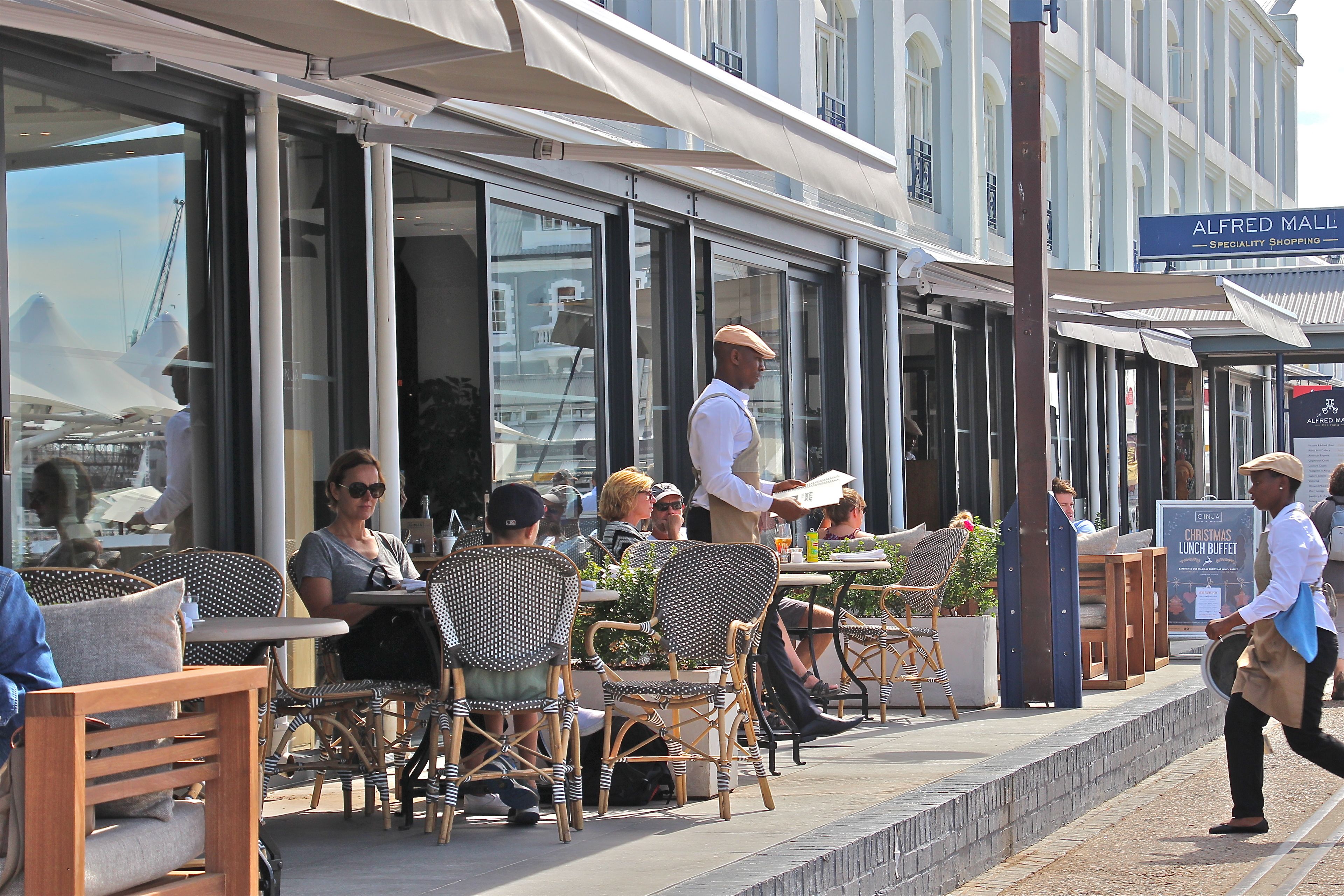 An outdoor cafe with wicker chairs and tables, a man walking by, and a sign for Alfred Mall.