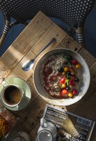 A wooden table with a bowl of cereal topped with berries and a spoon, a cup of coffee, and a magazine.