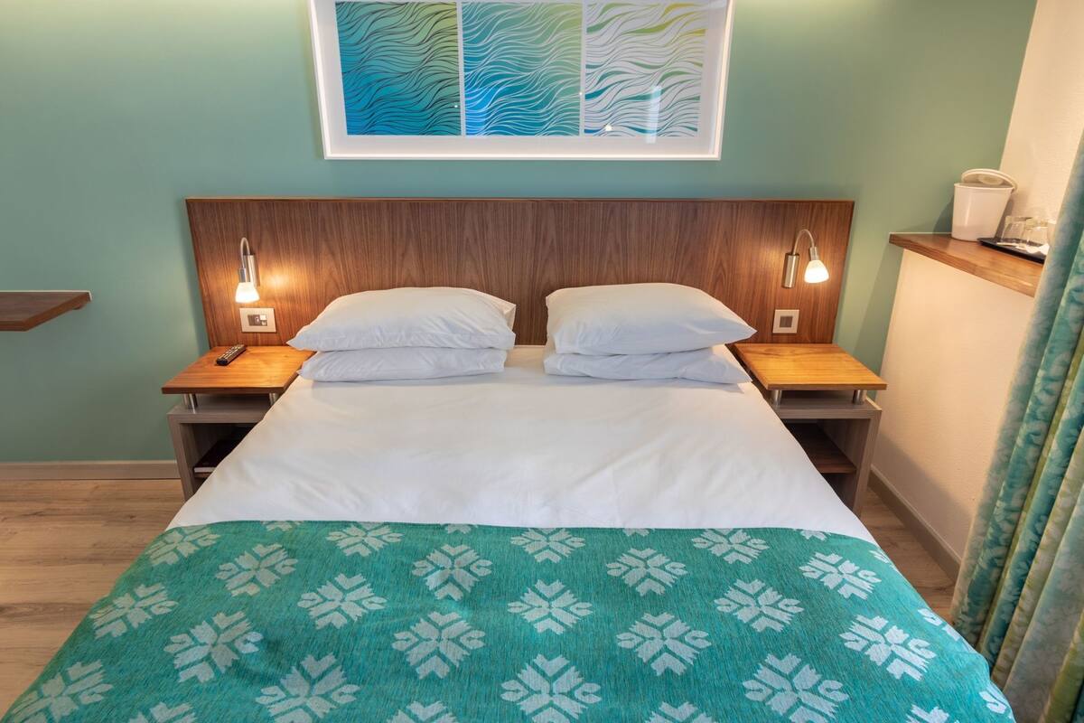 A double bed with a white duvet and a green patterned throw, two bedside tables with lamps, a wooden headboard, and a framed abstract artwork above the bed.