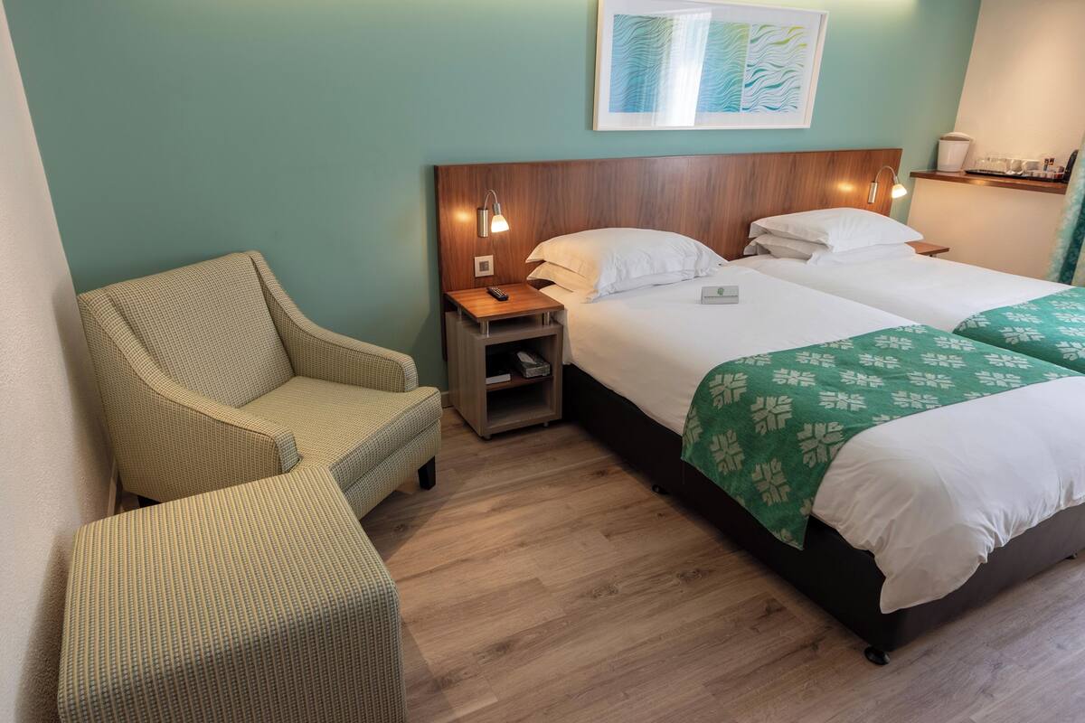 A hotel room with a wooden headboard, two beds with white linens, a green floral bedspread, a small nightstand with a lamp, a wicker chair, and a wall-mounted artwork.
