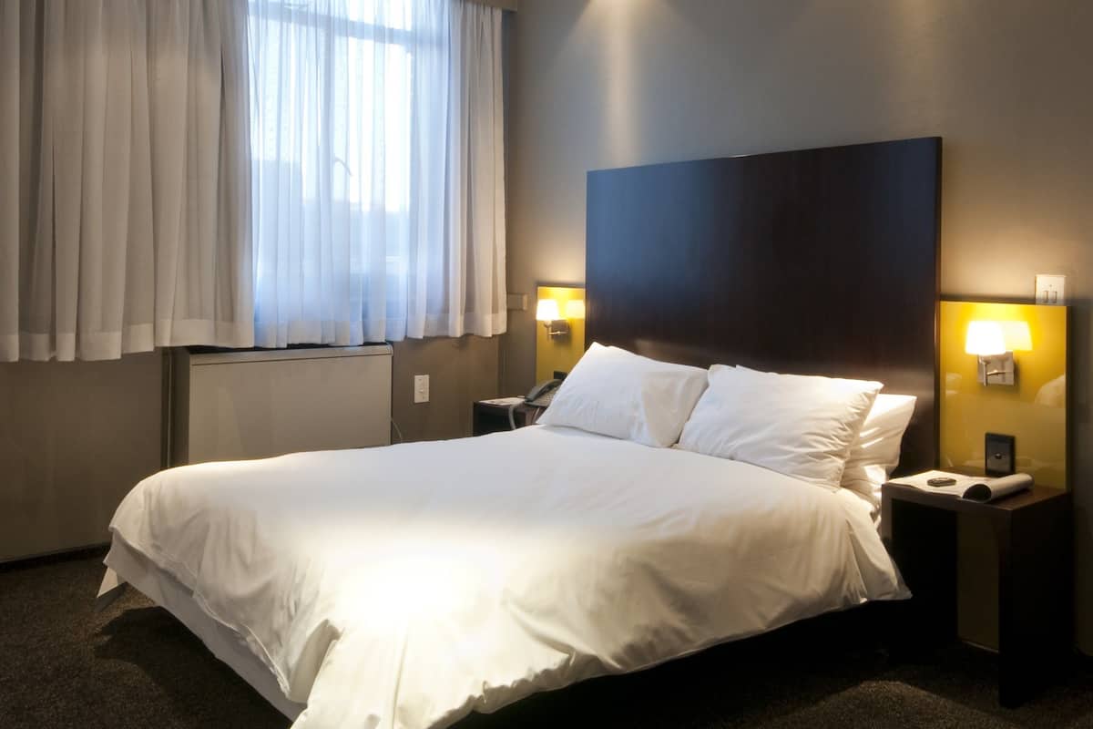 Une chambre d’hôtel avec un grand lit, deux tables de chevet avec des lampes, une fenêtre avec des rideaux légers et une tête de lit sombre.